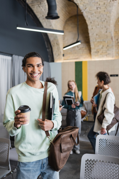 cheerful african american man with leather bag and coffee to go near students in blurred classroom - Photo, image