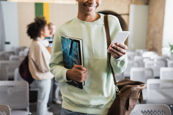 cropped view of african american man with magazine and notebook using smartphone on blurred background - Foto, Imagen