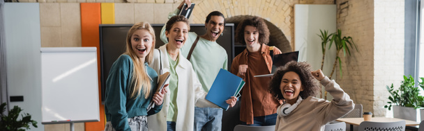 excited interracial students with copybooks showing win gesture in classroom, banner - Fotoğraf, Görsel
