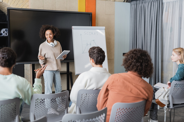 Afrikaans amerikaanse vrouw met digitale tablet in de buurt whiteboard met taak en interraciale studenten in de klas - Foto, afbeelding