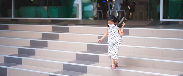 Προσωπογραφία παιδιού ηλικίας 5-6 ετών. Κοριτσάκι με λευκή μάσκα προσώπου για την πρόληψη της τοξικής σκόνης pm2.5 και του ιού. Χαριτωμένο παιδί έτρεχε προσεκτικά κάτω από τις σκάλες, κρατώντας τσάντα στο χέρι της. Κενό διάστημα. - Φωτογραφία, εικόνα