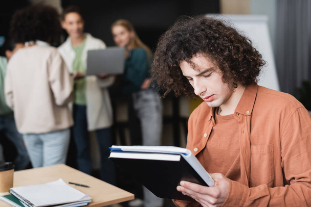 curly student with copybooks thinking near classmates talking on blurred background - Photo, Image