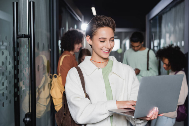 joyful woman using laptop while standing near interracial students in blurred corridor - Photo, Image