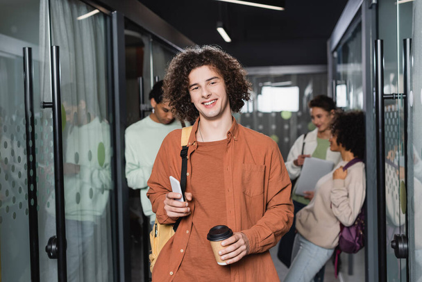 szczęśliwy młody człowiek z kawą i smartfonem patrzący na kamerę w pobliżu wielonarodowych studentów w zamglonym korytarzu - Zdjęcie, obraz
