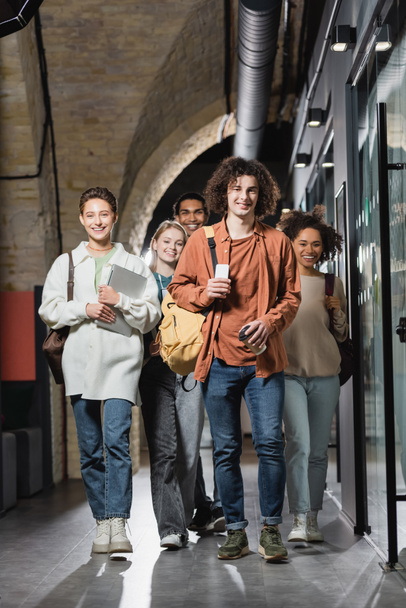 full length of smiling interracial students with gadgets walking along hallway in university - Valokuva, kuva