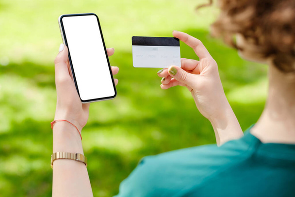 クレジットカードや電話を持っている赤毛の女性の手のビューを閉じて、モックアップ白い空白のディスプレイ、ショッピングや銀行アプリのための空の画面。モバイルアプリ技術の概念,肩の上に近いビュー. - 写真・画像