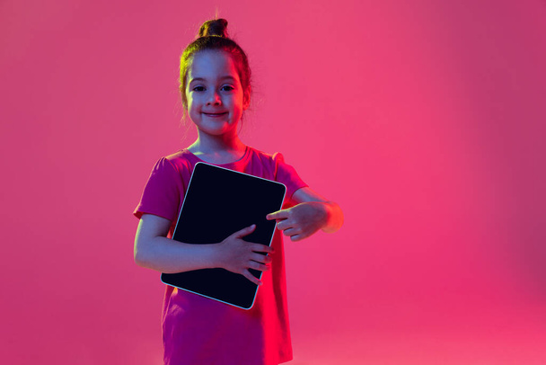 Pointing at gadget. Emotional kid, preschool age girl wearing pink t-shirt with digital tablet isolated on magenta color background. Concept of children emotions, fashion, beauty, education concept. - Foto, afbeelding