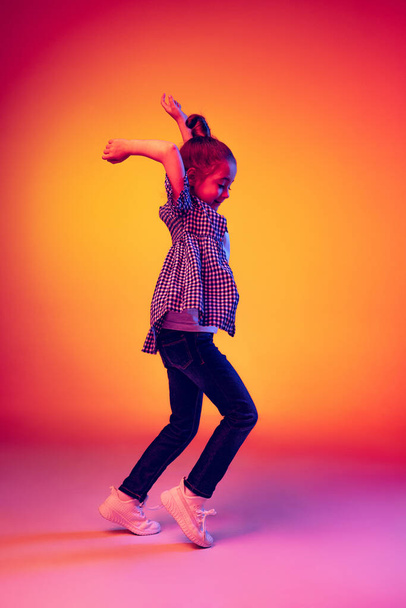 Jumping. Funny little girl, kid wearing warm plaid shirt having fun isolated on yellow red background in neon. Concept of children emotions, fashion, beauty, school and ad concept. Kids fashion - Photo, Image