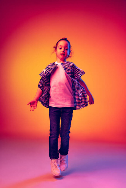 Jumping. Funny little girl, kid wearing warm plaid shirt having fun isolated on yellow red background in neon. Concept of children emotions, fashion, beauty, school and ad concept. Kids fashion - Fotó, kép
