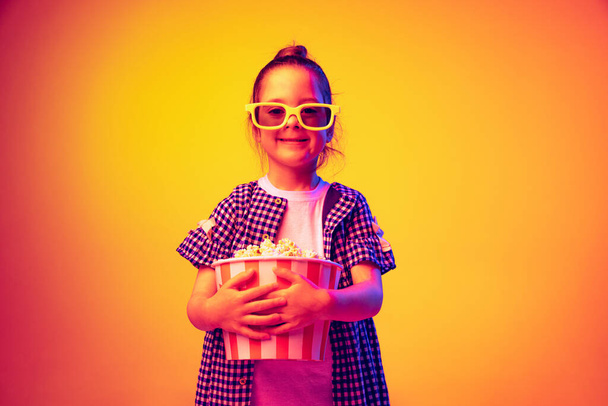 Watching comedy. One happy little girl, pupil wearing 3d glasses and holding bucket of popcorn isolated on orange background in neon. Concept of emotions, facial expression, youth, aspiration - 写真・画像