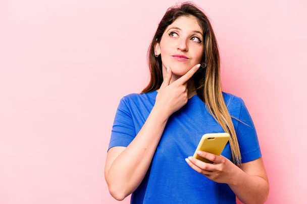 Молода біла жінка тримає мобільний телефон ізольовано на рожевому тлі, дивлячись убік з сумнівним і скептичним виразом
. - Фото, зображення