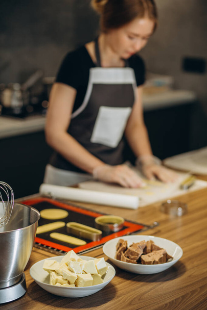 Mooie vrouwelijke banketbakker bereidt snoepjes in een moderne keuken. Chocolade in een bord op de voorgrond - Foto, afbeelding