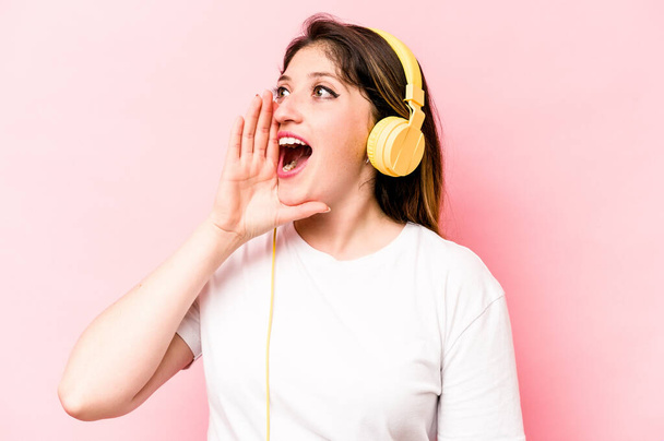 Young caucasian woman listening to music isolated on pink background shouting and holding palm near opened mouth. - Foto, imagen