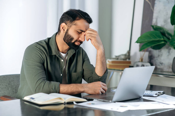 Tired exhausted arabic or indian man, office worker, manager or freelancer, sitting at his desk, tired of working in a laptop, overworked, having a headache, closed his eyes, needs rest and break - 写真・画像