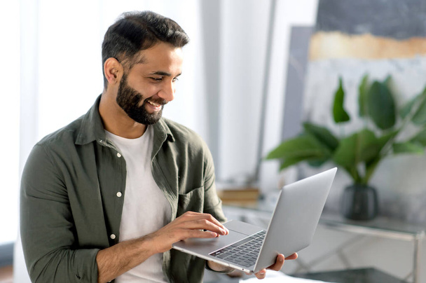 Happy attractive arabian or indian man, stock trader, real estate agent, holds laptop while stand in modern office, rejoices at the conclusion of a deal, high profits, profitable collaboration, smile - Foto, Imagen