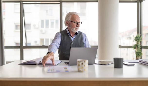 Серьезный бизнес-профессионал, работающий над ноутбуком. Задумчивый пожилой руководитель сидит за столом в офисе. Он носит формальности на работе.. - Фото, изображение