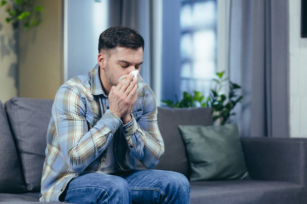 Feeling unwell. A young handsome man is sitting on the couch at home, is sick, feels cold, wipes his nose with a napkin, has a runny nose. - Foto, imagen