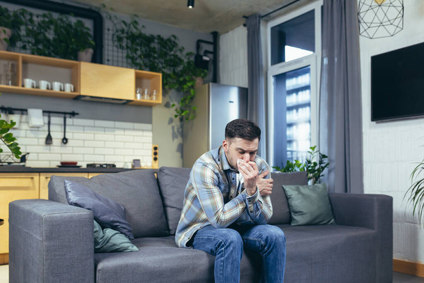 Feeling unwell. A young handsome man is sitting on the couch at home, is sick, feels cold, wipes his nose with a napkin, has a runny nose. - Fotoğraf, Görsel