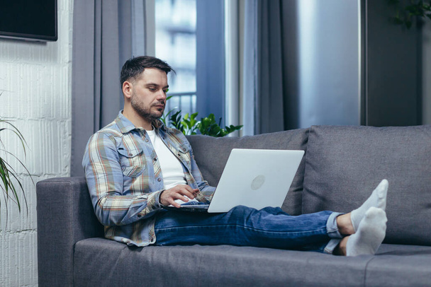 Freelancer. Handsome young man working on laptop at home. Lying on the couch in home clothes. - Photo, Image