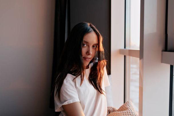 Close up portrait of european lovely woman with dark loose hair wearing white t-shirt posing at camera in sunlight near the window.  - Zdjęcie, obraz