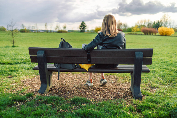 Mujer rubia caucásica de 30 años descansando, relajándose sentada en el banco y disfrutando en el verde parque natural de la ciudad al aire libre. Vista trasera  - Foto, imagen