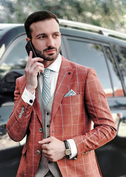 Young bearded man is talking on phone in tuxedo in front of car on street looking to side. - 写真・画像