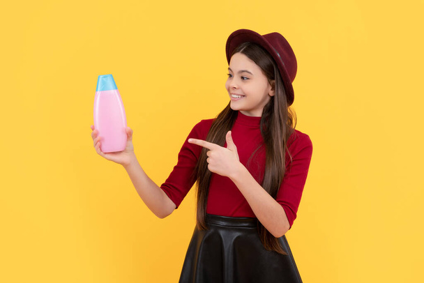 daily habits and personal care. happy girl in hat point finger on shampoo. presenting cosmetic product for teen. kid use shower gel. shampooing long hair in salon. child with conditioner. - Photo, Image