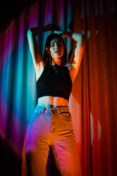 Young and sexy woman with wearing jeans and black t-shirt posing in colorful neon light - Фото, изображение