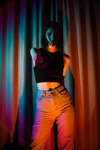 Jonge en sexy vrouw met jeans en zwart t-shirt poserend in kleurrijk neon licht - Foto, afbeelding