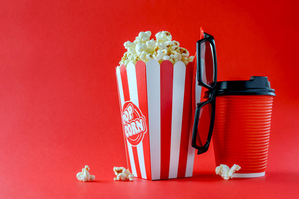 Popcorn in a striped cardboard box, 3d glasses, a paper glass with a drink on a red background, copy space. Movie Time. - Photo, Image