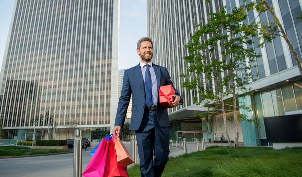 рад уверенный предприниматель в деловом костюме держа сумку для покупок и Giftbox за пределами офиса, случай. - Фото, изображение