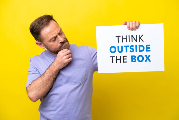 Middelbare leeftijd blanke man geïsoleerd op gele achtergrond met een bordje met tekst Denk buiten de doos en denken - Foto, afbeelding