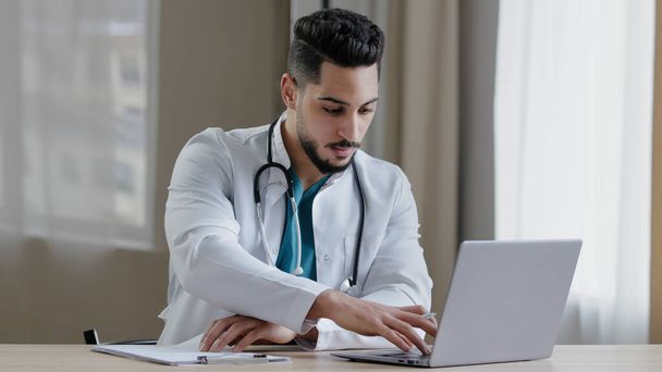Soustředěný arabský lékař praktický sedět na klinice kabinet použití počítače chatování vzdálený v lékařské sociální aplikace dát on-line konzultace psaní recept diagnostika vyhledávací informace - Fotografie, Obrázek