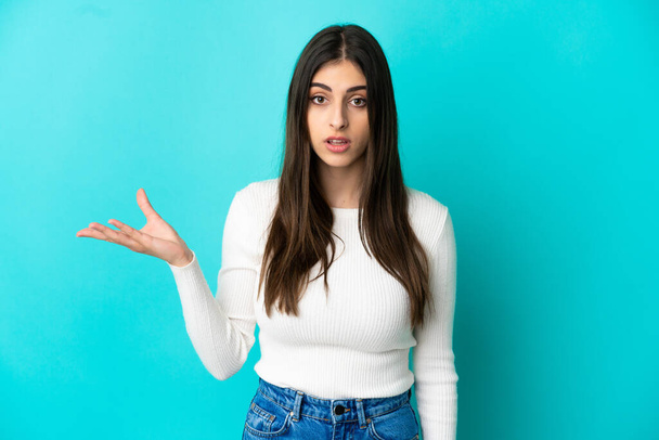 Young caucasian woman isolated on blue background making doubts gesture - 写真・画像