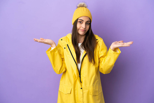 Young caucasian woman wearing a rainproof coat isolated on purple background having doubts while raising hands - Photo, Image