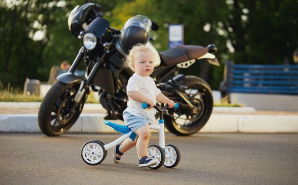 Cute baby learns to ride a balance bike. A small child tries to ride a bike in a sunny park with a big motorcycle in the background. The kid dreams of becoming a biker - Photo, Image