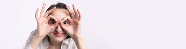 Крупный план портрета привлекательной причудливой молодой женщины, делающей бинокль руками, показывающими жест ОК на белом фоне студии. - Фото, изображение