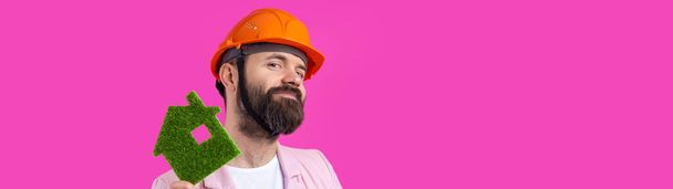 Portrait of young construction engineer wear orange hard hat, in a pink jacket standing on red studio background. A man holds a green eco house. - Φωτογραφία, εικόνα