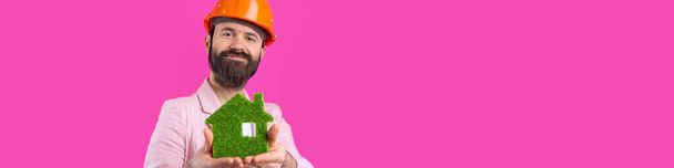 若い建設エンジニアの肖像画は、赤いスタジオの背景に立ってピンクのジャケットで、オレンジのハード帽子を着用してください。男は緑のエコハウスを持っている. - 写真・画像