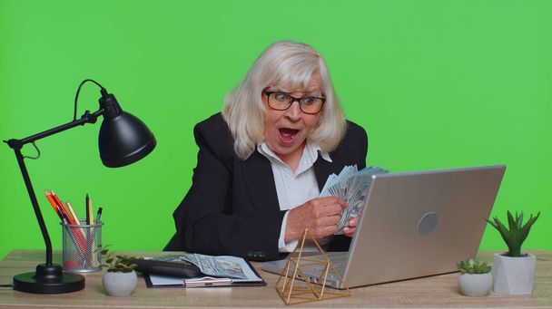 Ανώτερη λογίστρια επιχειρήσεων ή τραπεζίτης κάνει υπολογισμούς στο γραφείο στο χώρο εργασίας. Ηλικιωμένη γιαγιά καταμέτρηση των εσόδων σε μετρητά δολαρίων, που εργάζονται στο γραφείο στο πράσινο στούντιο κλειδί chroma - Φωτογραφία, εικόνα