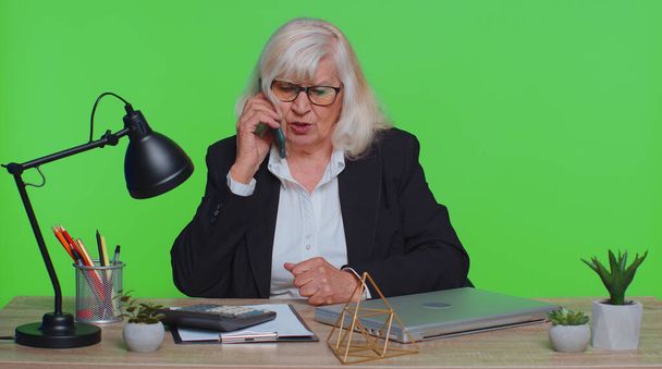 Wütende ältere Geschäftsfrau, die am grünen Büroarbeitsplatz telefoniert, genervt ceo sprechende Handygespräche verärgert wütend. Arbeiterinnen-Chef-Handy Streit Probleme negative Emotionen - Foto, Bild