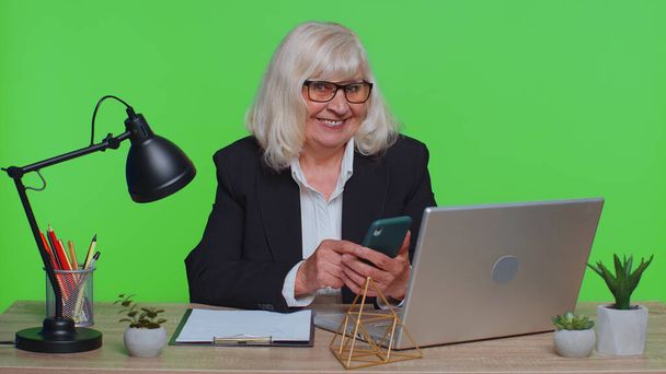 Зріла бізнес-леді, використовуючи мобільний телефон, вводить новий пост на веб смс-повідомлення, перегляд, пошук. Залежність від соціальних мереж в офісі. Старша бабуся студійний знімок ізольовано на зеленому хромовому ключі
 - Фото, зображення