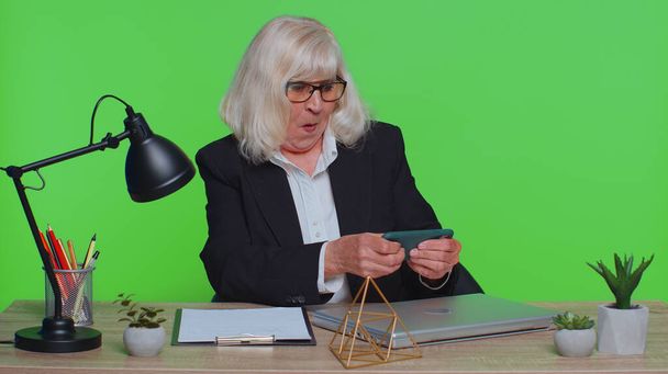 Worried funny senior businesswoman sits at workplace avoid working and enthusiastically playing racing or shooter video games at office desk table. Elderly woman on green chroma key studio background - Fotografie, Obrázek