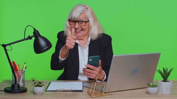 Szczęśliwy podekscytowany radosny starszy businesswoman używać smartfona wpisując przeglądanie powiedzieć wow tak, wielkie zwycięstwo dobre wieści zszokowany nagłym zwycięstwem w biurze. Starsza kobieta odizolowana na studiu zielonego klucza chroma - Zdjęcie, obraz