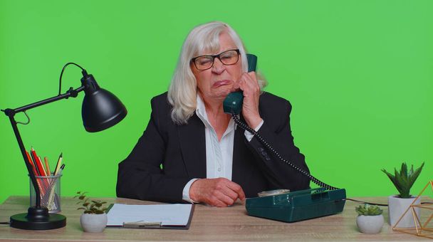 Crazy senior businesswoman talking on wired vintage telephone of 80s, fooling, making silly faces, humor comic, sits at workplace desk at office. Elderly woman isolated on green chroma key studio - Photo, image