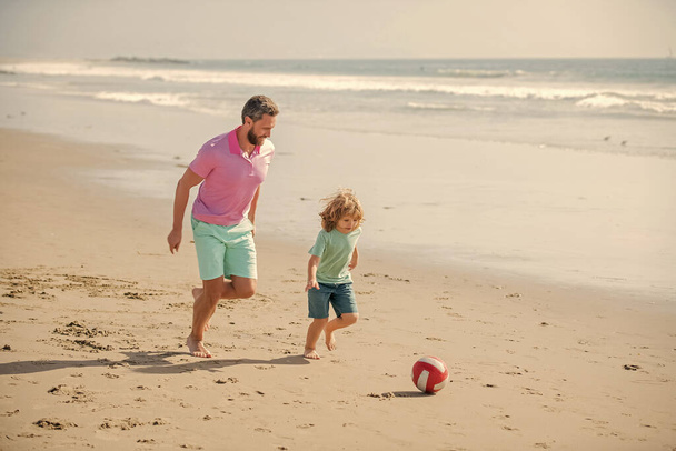 Παιδική ηλικία και ανατροφή. οικογενειακές διακοπές. αθλητική δραστηριότητα. Πατέρας και γιος παίζουν ποδόσφαιρο στην παραλία. Ο μπαμπάς με τον μικρό σε μια αστεία καλοκαιρινή μέρα. Ημέρα της οικογένειας το Σαββατοκύριακο. μπαμπάς και παιδί διασκεδάζουν έξω.. - Φωτογραφία, εικόνα