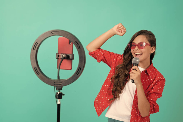 szczęśliwy nastolatek dziewczyna piosenkarka w okularach przeciwsłonecznych śpiewa piosenkę na aparat wideo blog blogowanie online z selfie led i mikrofon, karaoke online. - Zdjęcie, obraz