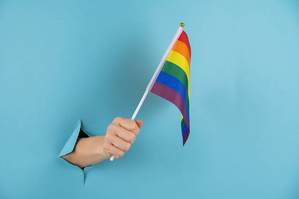 A womans hand sticks out of a hole in a blue paper background and holds an lgbt flag - 写真・画像
