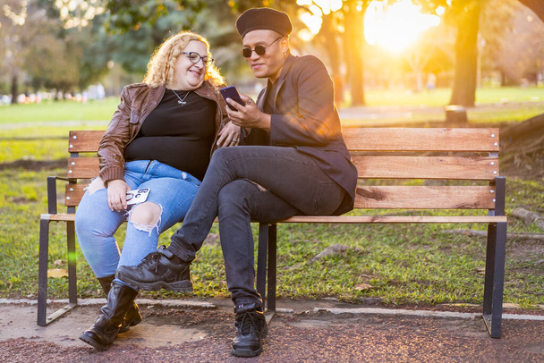 Gay přátelé sedí na lavičce v parku při západu slunce a povídají si a dívají se na mobil - Fotografie, Obrázek