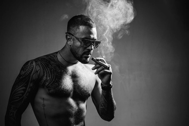 Tattooed man with cigar. Cigar smoking enjoy life and moment. Portrait of a bearded businessman with a beautiful torso smoking a cigar and drinking whiskey. - Foto, Imagen
